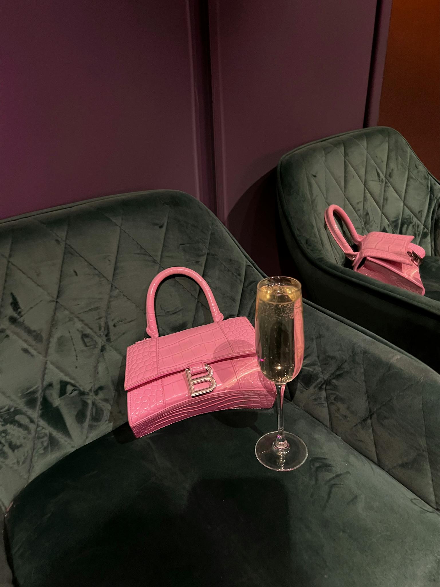 Elegant pink handbag with champagne glass on plush chair in a stylish setting.